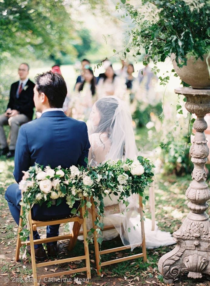 Groom and bridesit on chairs with flowers seen from behind